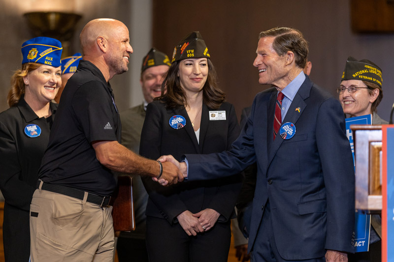 A veteran wearing a black polo shirt and khakis shakes the hand of Senator Richard Blumenthal at an event as others in uniform stand nearby watching while smiling. 