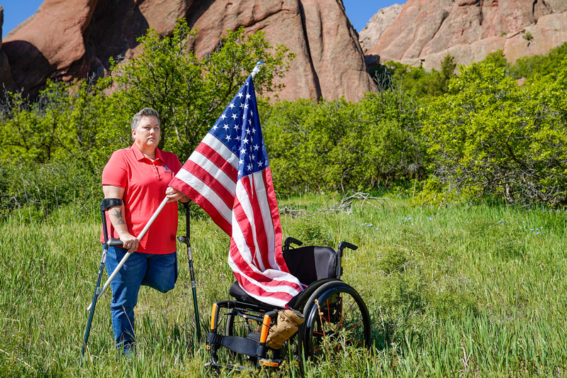 Female veteran leg amputee standing with crutches in a grassy field beside a wheelchair holding an American flag. 